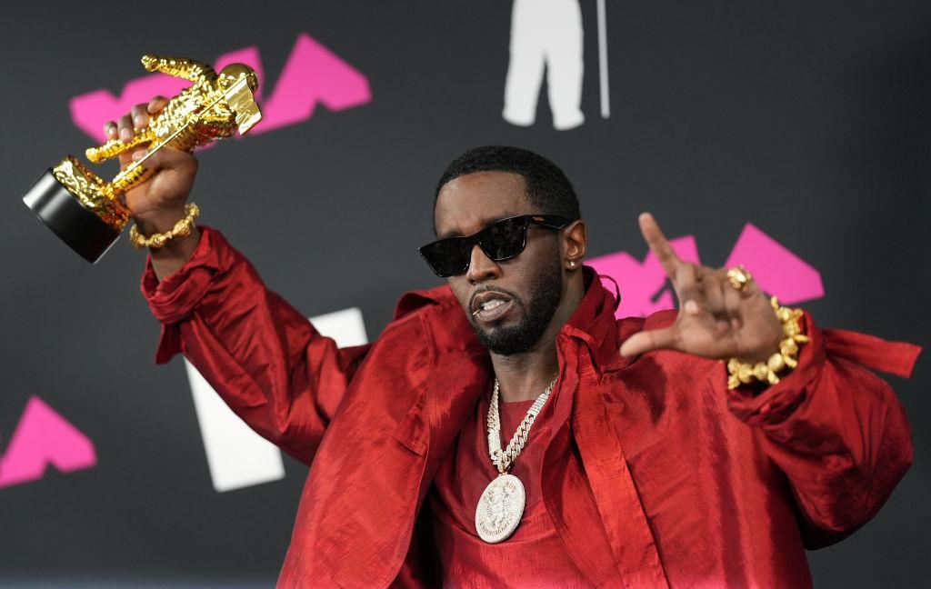 Diddy holds up a VMA award on the red carpet and wears a red linen jacket and shirt and a large silver Bad Boy chain.