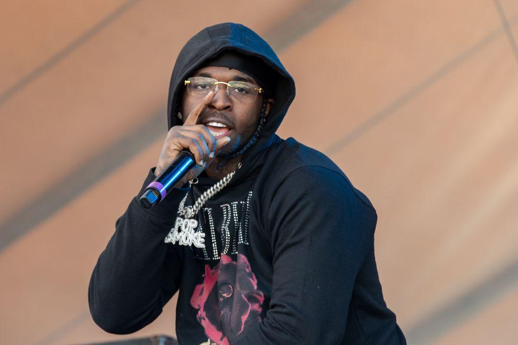 Pop Smoke holds a microphone at performs on stage wearing a black and redd bedazzled hoodie.