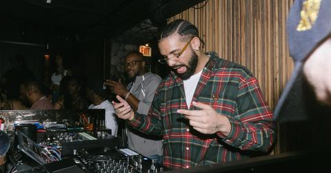 Drake wearing a flannel button down, white t-shirt, and stands behind a DJ booth.