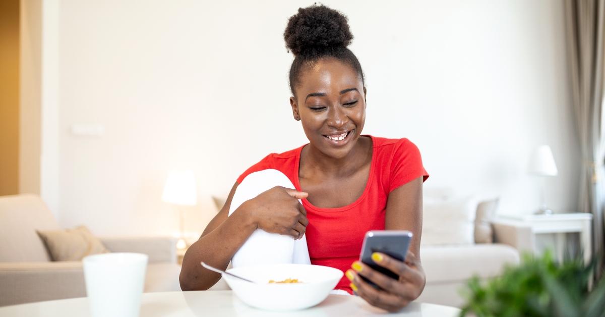 Young Black woman eats breakfast at a table while smiling at her phone.
