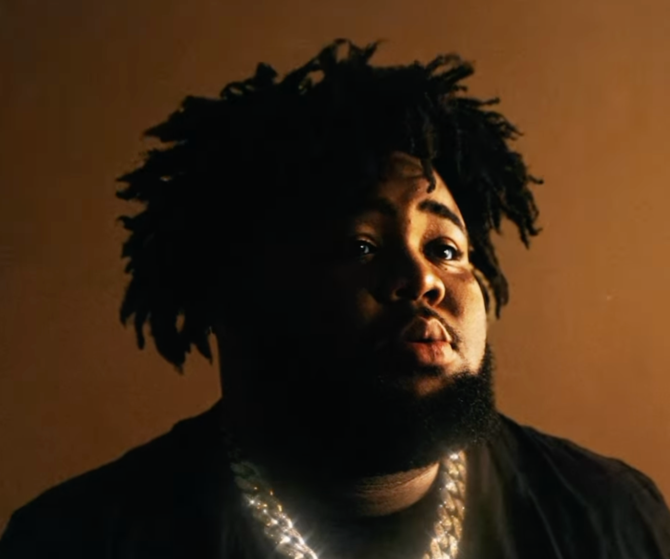  Rod Wave wearing a gold chain and a black shirt against brown background.