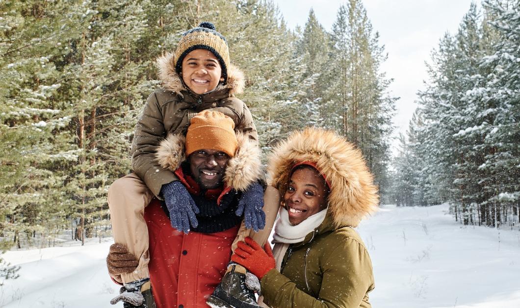 Cheerful family of three in warm winterwear spending day in winter forest or park among firtrees and pines covered with snow.