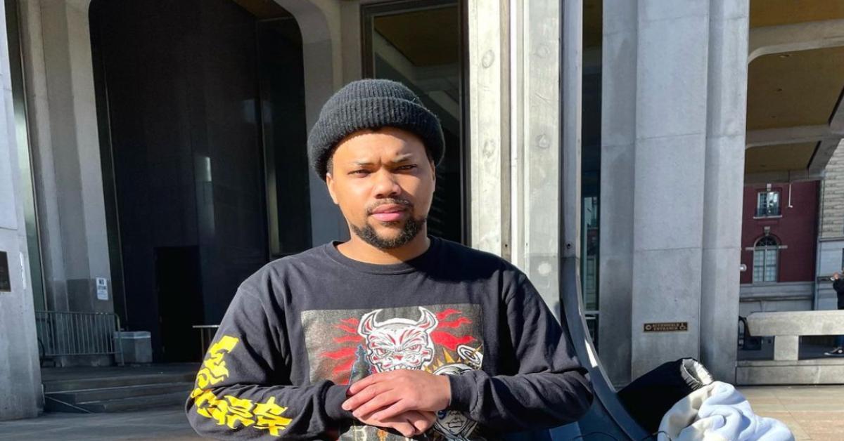 Rapper QThree stands near concrete steps wearing black shirt and beanie.