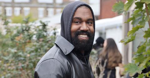 Kanye West wears a black hoodie and leather jacket and smiles at paparazzi in London.