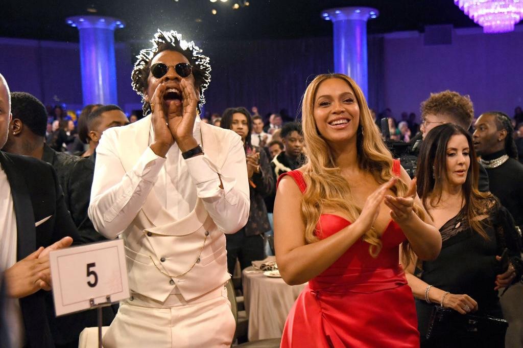 Jay Z wears a white tuxedo and cheers standing next to Beyoncé smiling and wearing a red dress.