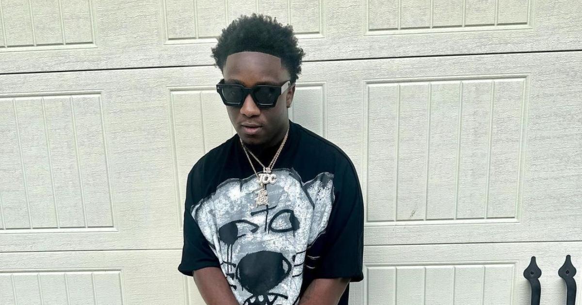 Lil Joc poses next to white garage wearing a black shirt, sunglasses, and a chain.