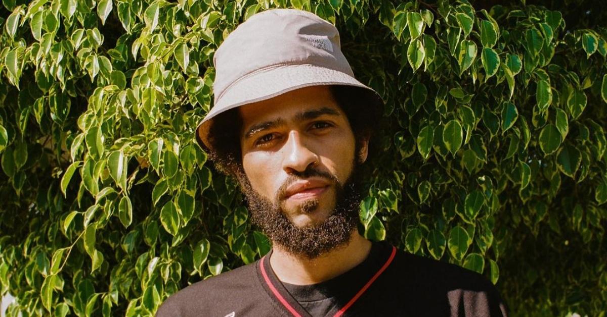 Rapper Lojii stands by a tree and wears a bucket hat and jersey.