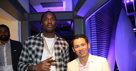 Meek Mill wears a white t-shirt, plaid button down shirt, and gold chains and holds up the piece sign. Johnny Dang wears a grill and a yellow suit and smiles.
