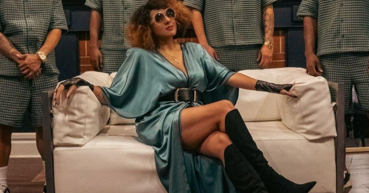 Marsha Ambrosius poses while seated on a couch with men in matching outfits standing behind her