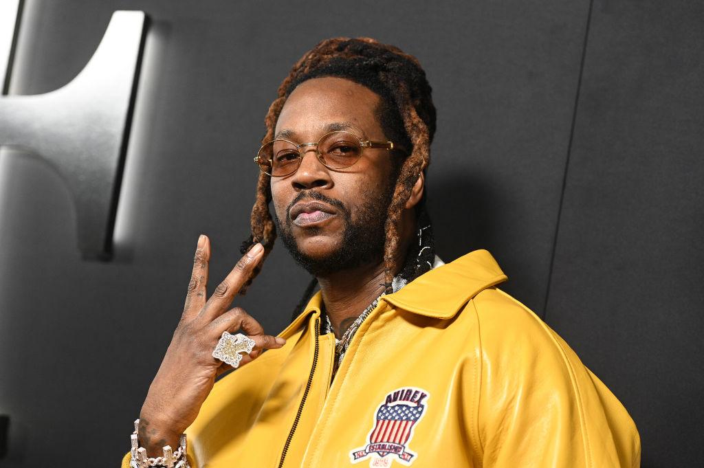 2 Chainz holds up a peace sign and wears a yellow leather Avirex jacket and diamond ring.