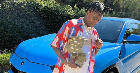 Soulja Boy wears chains and holds a stack of money standing by a blue sportscar.