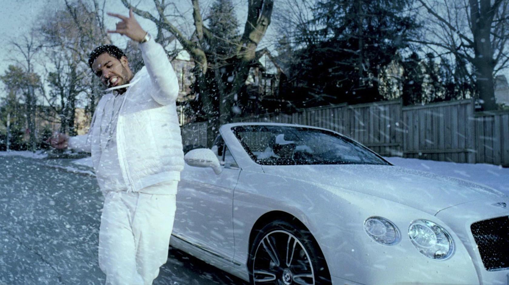 Drake dances in the snow wearing an all white outfit next to a white convertible.