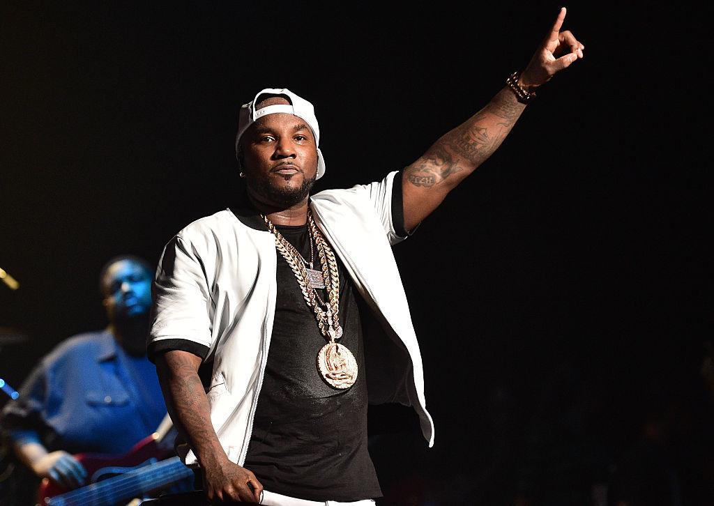 Jeezy stands on stage with one finger in the air wearing a gold chain and black shirt.