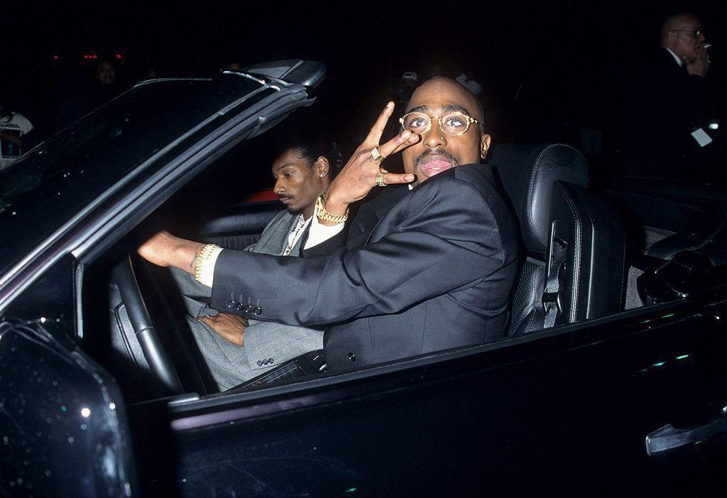 Tupac holds up the west coast hand gesture while wearing a tuxedo in the driver's seat next to Snoop Dogg.