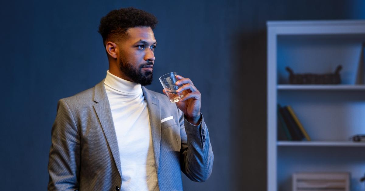 A young african american businessman with glass of water indoors in office at night, looking aside.