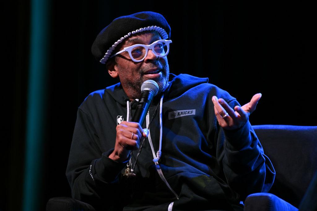 Spike Lee wears a black Knicks Nike tracksuit and a black beret and holds microphone and speaks.