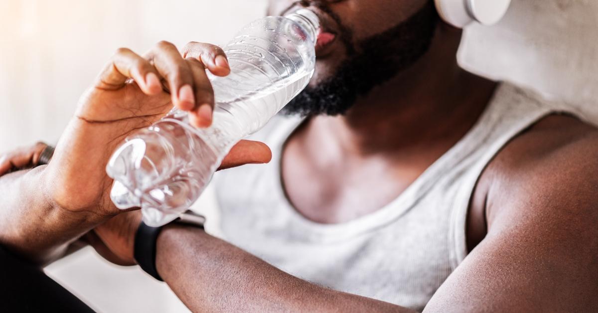Close up of hands of young bearded black athlete holding bottle of water. He is sitting on floor, leaning on wall and drinking