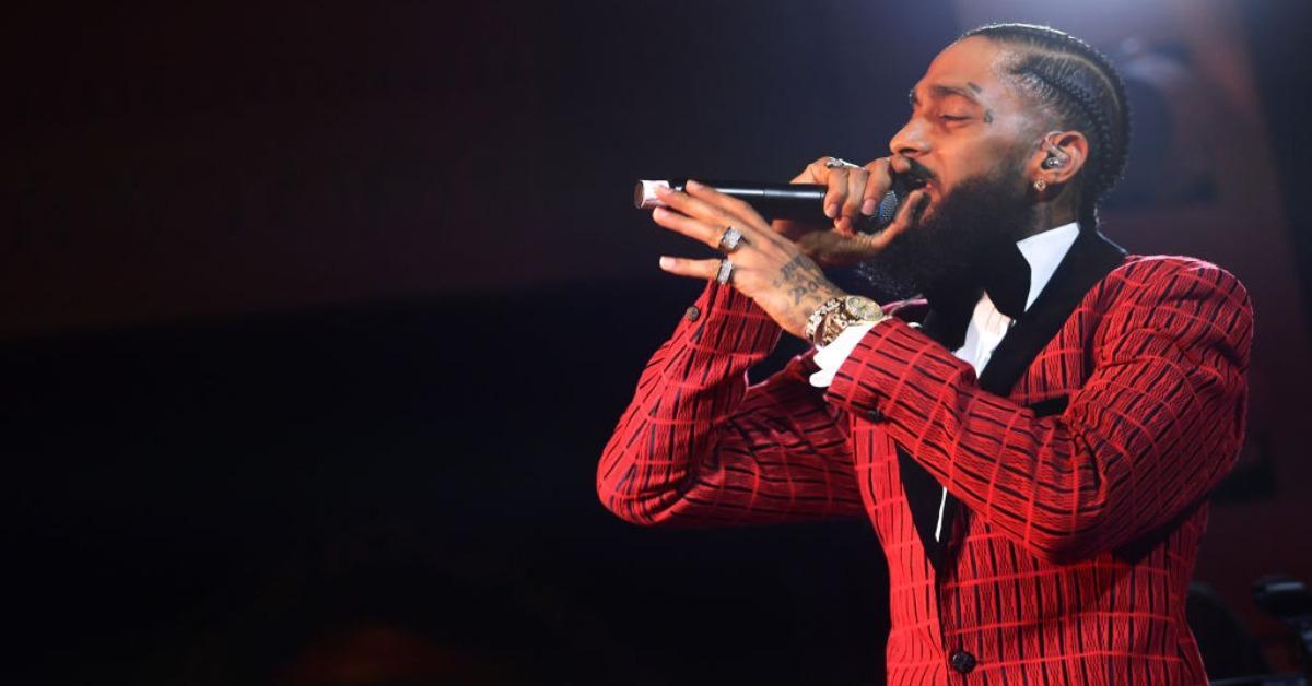 Nipsey Hussle raps on stage at the Warner Music Pre-Grammy Party in 2019 wearing red suit jacket.