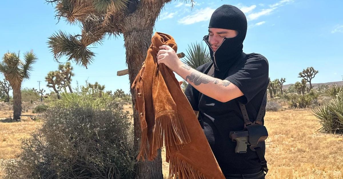 Year wears a black zip-up ski mask and holds a brown jacket standing in the desert.