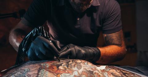 Black man bent over tattooing a colorful tattoo on a man at a tattoo shop.