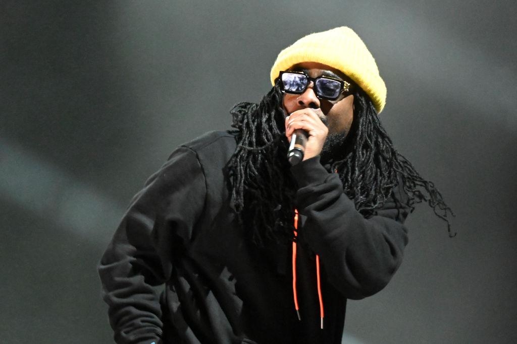 Wale performs on stage wearing a black jacket and a beanie.