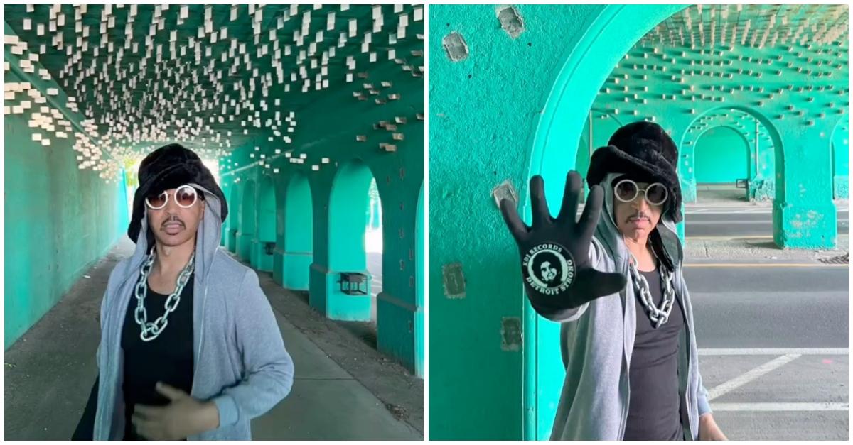 Moodyman poses in a blue corridor wearing silver chain link, a grey hoodie, black hat, and rhinestone-studded glasses.