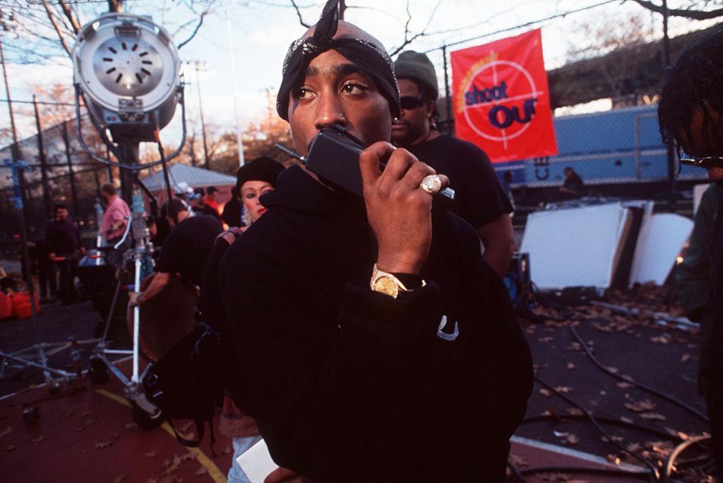 Tupac wears a black bandana and hoodie and talks into a walkie talkie on the set of a movie.