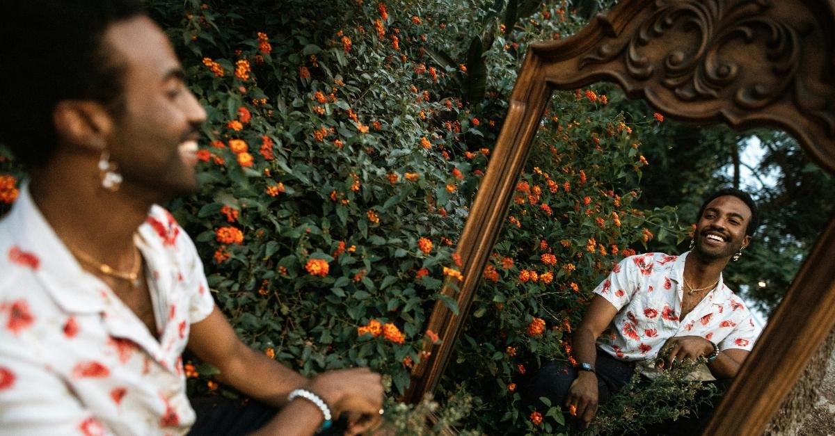 A black man smiles and looks into a mirror while sitting in a field of flowers.